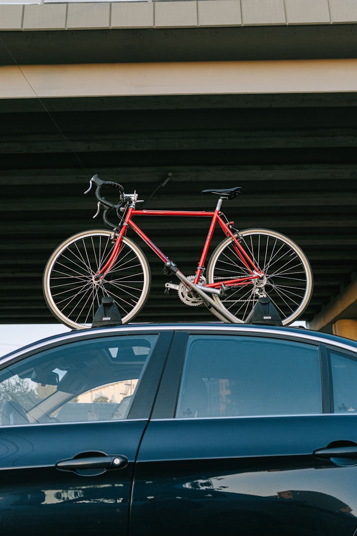 Red road bike securely mounted on a car roof rack under a bridge.
